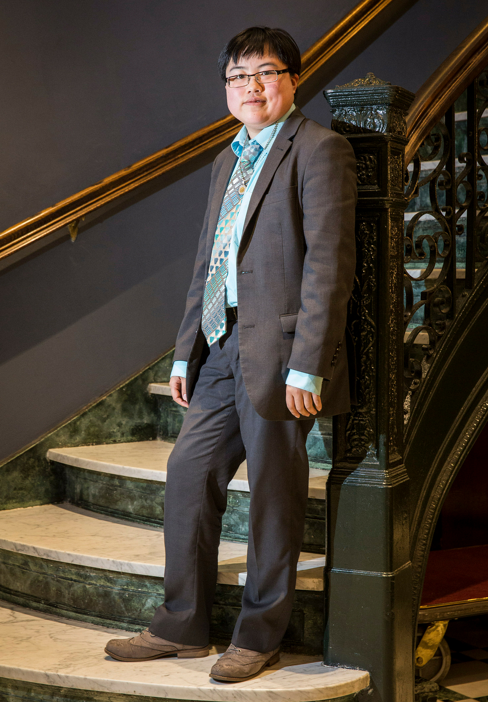 Photo: Lydia Brown, a young East Asian person with short black hair and glasses, standing against a wrought-iron banister and marble staircase in a darkly painted room. Lydia has a serious expression and is wearing a gray suit with teal shirt and tie.